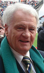 An elderly man with grey hair smiling at the camera. He is in a football stadium and is wearing a black suit with a multi-coloured tie and a green scarf.