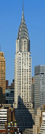 Distant ground-level view of an 80-story building; the structure has a stone, whitish exterior with several setbacks. Statues project from the building near the 30th and 60th floors, and the building tapers into a thin, silver spire containing angled triangular windows.