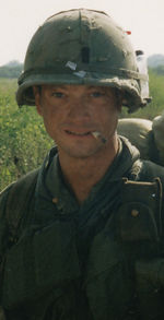 A man is at the center of the image looking at the camera. He is dressed in Vietnam-era military attire including a vest and helmet. He has a cigarette sitting on his lips and is wearing a backpack.