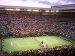 A green tennis hard court, in the middle of a stadium with crowded bleachers, under a blue sky