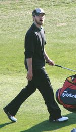 A brunette man with facial hair walks over grass holding a golf club.