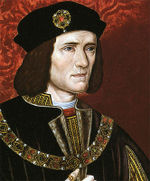 A man with furrowed brows, cleft chin, and shoulder length black hair. He is dressed in black, with a thick jewelled chain and a crest on his hat.