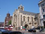 A street sloping uphill from right to left. There are parked cars and pedestrians in the foreground, and the hoardings further up indicate that the road is temporarily closed. On the right of the road in the centre of the picture is one end of a large early twentieth-century stone-built church in the Arts and Crafts style which is apparently being restored, as there are portakabins and screens outside. It has an elaborate entrance on the street with a large recessed window above and another to the right. There is another, plainer, entrance on the minor street with a No Entry sign that runs alongside the side of the church. Alongside it are two small windows; above and behind this entrance and the windows are three large arches with, underneath each, a tall thin window. Beyond these, part of a transept can be seen. Above the church is a bell-tower with castellated columns at the corners and a pyramid roof. There are railings between the church and pavement. Beyond the church, some red-brick buildings can be seen, most notably a four-storey building with an elaborate corner tower. In the right foreground, across the side-street from the church, part of a four-storey 1920s neo-Georgian building can be seen behind a pair of telephone kiosks.