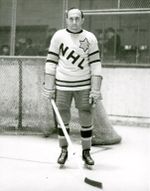 An ice hockey player stands on an ice rink. He is wearing similar equipment as before, except that his sweater has the letters "NHL" in a downward diagonal with a large star on his left shoulder.