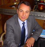 A seated man with dark hair looks up to the camera. He is wearing a grey pinstripe jacket, light blue shirt, dark blue tie, and a ring on the fourth finger of his left hand. There is a commputer keyboard to his left.