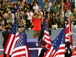 A man in brown shorts and an orange shirt holds a silver trophy, with photographers shooting him in the background, and several American flags in the foreground