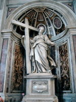 A marble statue showing a matronly woman in a sweeping cloak supporting a cross which stands beside her and gesturing to the viewer with her left hand