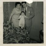 A woman and man, both in their thirties and both dressed in 'Sunday best', hold a similarly dressed very young girl standing on the arm of a floral-print sofa.