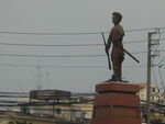 Side on view of a cement statue depicting a man with a moustache holding a  rifle in one hand and a stick in another, wearing traditional clothes. He stands on a polished stone pedestal, and a city landscape and buildings can be seen in the background.