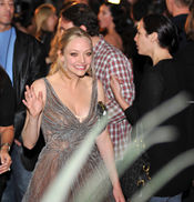 A young blonde female wearing her hair in a ponytail and a silver low-cut dress waving and smiling.