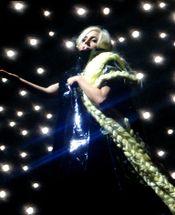 A blond woman wearing a shiny black dress and her hair extended with long braids, stands on a stage. The braids fall on her left arm and continue down. Behind her, a number of twinkling lights are visible and the woman makes a gesture with her right hand.