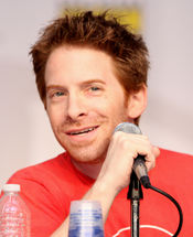 A man with red hair, smiling slightly and sitting behind a microphone.