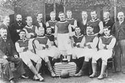 A black and white photograph showing two rows of people in front of a building; the front row are seated with a trophy in the middle on a box covered in a striped cloth; the rear row are standing.