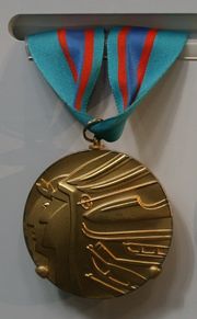 Close-up of a golden medal with a stylized embossed design of two overlaping faces in profile. One is wearing a laurel wreath and the other a native American headdress, whose feathers are represented by several winter sports equipments and gear. The medal is suspended in a red and light blue ribbon.