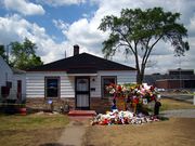 A house surrounded by yellow colored grass, flowers, trees, and a light blue colored sky can be seen. The house has white walls, two windows, a white door with a black door frame, and a black roof. In front of the house there is a walk way, yellow grass and multiple colored flowers and memorabilia. In the background, there are two tall trees and a light blue colored sky that has multiple clouds.