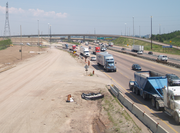 "Construction alongside a major freeway. In the background is a large interchange. Buildings are visible to either side of the freeway."