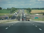 A highway rolls over hills as it climbs into the distance against an evening sky. The highway is eight lanes wide with considerable traffic flow. In the distance, high-tension wire pylons arc across the view from left to right.