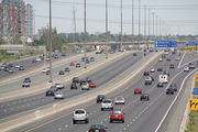 "A view of a wide freeway from the side. The freeway is divided into four segments, each of which contains three lanes (four in some places). The vehicles within the two sections nearest the camera are travelling away into the distance, whereas the vehicles within two sections further from the camera are approaching. Several signs dot the right side of the freeway, and two overhead gantries (also holding signs) are also visible."