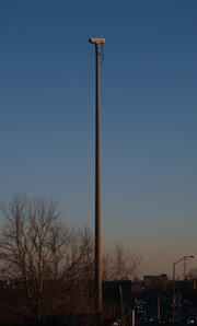 A video camera mounted on a tall cement pole on the side of a roadway. The camera is not pointing at the roadway visible at the bottom-right of the picture, but to the left.