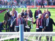 In the foreground is two men holding a large cup, they have claret scarves and a medal around their necks. Around them are ten old players in suits with medals and scarves around their necks.