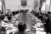 Twenty people meet in a conference room around an oval table. One man, at the center of the table on the right-hand side, is addressing the others. All are wearing suits or similar attire.