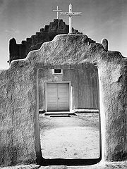 A black-and-white vertical photograph shows an adobe wall in the foreground, rising in the middle with a stair-step pattern and a white wooden cross at the pinnacle, with an open doorway beneath. Through the doorway and above the wall, an adobe church with white double doors and a similar stair-stepped roof and cross stands, slightly larger than the wall in front of it. The midday sun casts harsh shadows on the dirt ground.