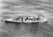 An aerial photograph of a large, World War I-era warship armed with four gun turrets, each with two guns, at sea. The ship is listing to port, but does not appear to be otherwise moving.