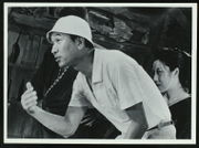 Kurosawa in early middle age wearing modern clothing is standing, or rather crouching, in the foreground of a dilapidated-looking studio set, gesturing with his hand and apparently talking to someone beyond the left edge of the frame; slightly behind him and to the right is the actress Isuzu Yamada, an attractive middle-aged woman in traditional Japanese garb.