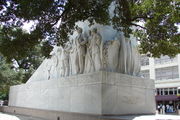 The rectangular base of a cenotaph. An angel is carved on one end. On the side are carvings of several men, shown wearing bucksin or 19th-century suits. Many hold guns or knives; at the far end, one operates a cannon.