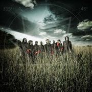 A group of nine men wearing masks stand in a field of grass with a gloomy clouded sky behind them.