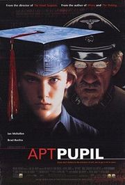 A teenage boy (played by Brad Renfro) stands on the left half of the image, seen from shoulder up.  He wears a collared blue shirt and a graduation cap (a square board on a skullcap that comes down near his eyebrows, all blue).  From the cap hangs a red tassel off the right side.  The left half of boy's face is lit up by a light source from off the right side of the image.  On the right side of the image stands an old man (played by Ian McKellen) immediately behind the boy's left shoulder, also seen from shoulders up.  He has round glasses and a white-and-gray goatee.  He is cast in shadow, and he wears a Nazi uniform (a gray coat with a black decorated collar, and a gray hat with a black visor and an emblem of an eagle on the cap above the visor).  The image's background is black, and the blackness slightly envelops the edges of the men's appearances.  Below their shoulders is the title "Apt Pupil" in uppercase.  "Apt" is colored red, and "Pupil" is colored white and is bolder than the previous word.