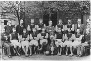 A black and white photograph showing two rows of people in front of a building; the front row are seated with two trophies in the middle, the rear row standing.