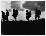 Five soldiers silhouetted against the sky. Rays of sun burst through dark clouds to create a dramatic and atmospheric shot. They are all wearing steel helmets, and three of them are clearly carrying rifles and backpacks.