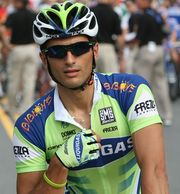 A cyclist wearing a green and blue jersey with white trim, a matching helmet and glove on his right hand (the only one visible), and sunglasses. His right hand hovers around the zipper on the front of his jersey, and vaguely visible behind him are three people in black shirts and khaki pants.