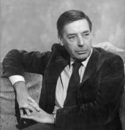 Half-length portrait of a man in his fifties with dark hair and a serious expression. He wears a dark sportcoat, light shirt, and dark tie, and his right arm is slung over the back of the sofa he is sitting on.