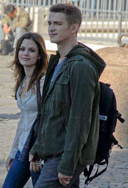 A woman and man, both smiling, are walking towards the left side of the image. The woman is wearing a white, button-up shirt and blue jeans. The man is wearing a green jacket, a blue shirt, a backpack, and blue jeans. In the background a man can be seen sitting and looking down.