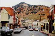 A steeply inclined road winds through tightly packed historical structures.  Several vehicles are parked in parallel formation on the left side of the road.  Signs advertise businesses on either side of the roadway, and electrical lines cross overhead.  In the background, a red colored hill rises against an overcast backdrop.