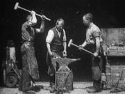 In a black-and-white screenshot, three men are standing around an anvil. The man on the left is lifting his sledgehammer above his head with both hands about to strike the anvil. The man in the center has a hammer in his right hand and is resting it on the anvil. The man on the right is holding a sledgehammer with both hands and is currently lifting it. The men are wearing blacksmith aprons, long sleeve shirts, and pants.