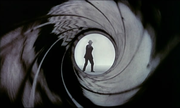 A man is seen through The barrel of a weapon, with the rifling pointing outside to a man wearing a business suit and hat, who points a gun at the viewer. The smoke around the man's face suggests he already fired.