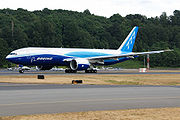 Side view of aircraft on runway. Runway surfaces in foreground and forest in background.