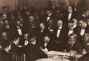 Three formally attired men at a conference table sign documents while 32 others look on.