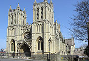 A stone built Victorian Gothic building with two square towers and a central arched entrance underneath a circular ornate window. A Victorian street lamp stands in front of the building and on the right part of a leafless tree, with blues skies behind.