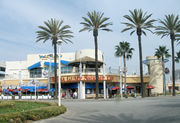 A two-story building has the sign "Bubba Gump Shrimp Co" on the front. Several people are in front of the building, and in the foreground is a paved road with some flowers on the left. Palm trees are located in front of the building and in the background there is blue, slightly cloudy skies.