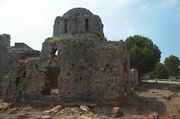 Ruins of a small stone domed structure built in a Byzantine style with tall windows. Grasses grow on the second level, as do trees behind it.