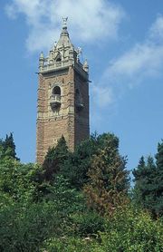 An ornate brick tower surrounded by trees. The tower has balconies and is surmounted by a pitched roof with an ornate figure at the apex.