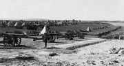 Foreground, a battery of 16 heavy guns. Background, conical tents and support vehicles.