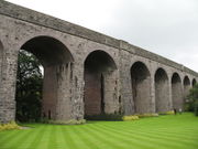 A series of seven stone archways, connected to stone parapet above grass area.