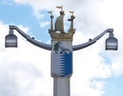 Post holding two lamps at the ends of two arms. The top of the post is decorated by a galleon; underneath the boat is a crest containing a white lower-left and a blue lower-right separated by a jagged line, below a white bird carring an olive branch.