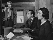 A stern looking man and a woman sit on the right side of a table with documents on the table. A top hat is on the table. An unkempt man stands to the left of the picture. In the background a boy can be seen through a window playing in the snow.