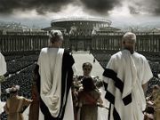 Several men in white robes are facing away from the image, at the top of large steps. A man is at the center of the image being handed flowers by a girl. In the background are rows of thousands of soldiers and members of a large crowd. In the distance, the Colosseum can be seen along with other buildings in Rome. Dark clouds are visible in the sky.